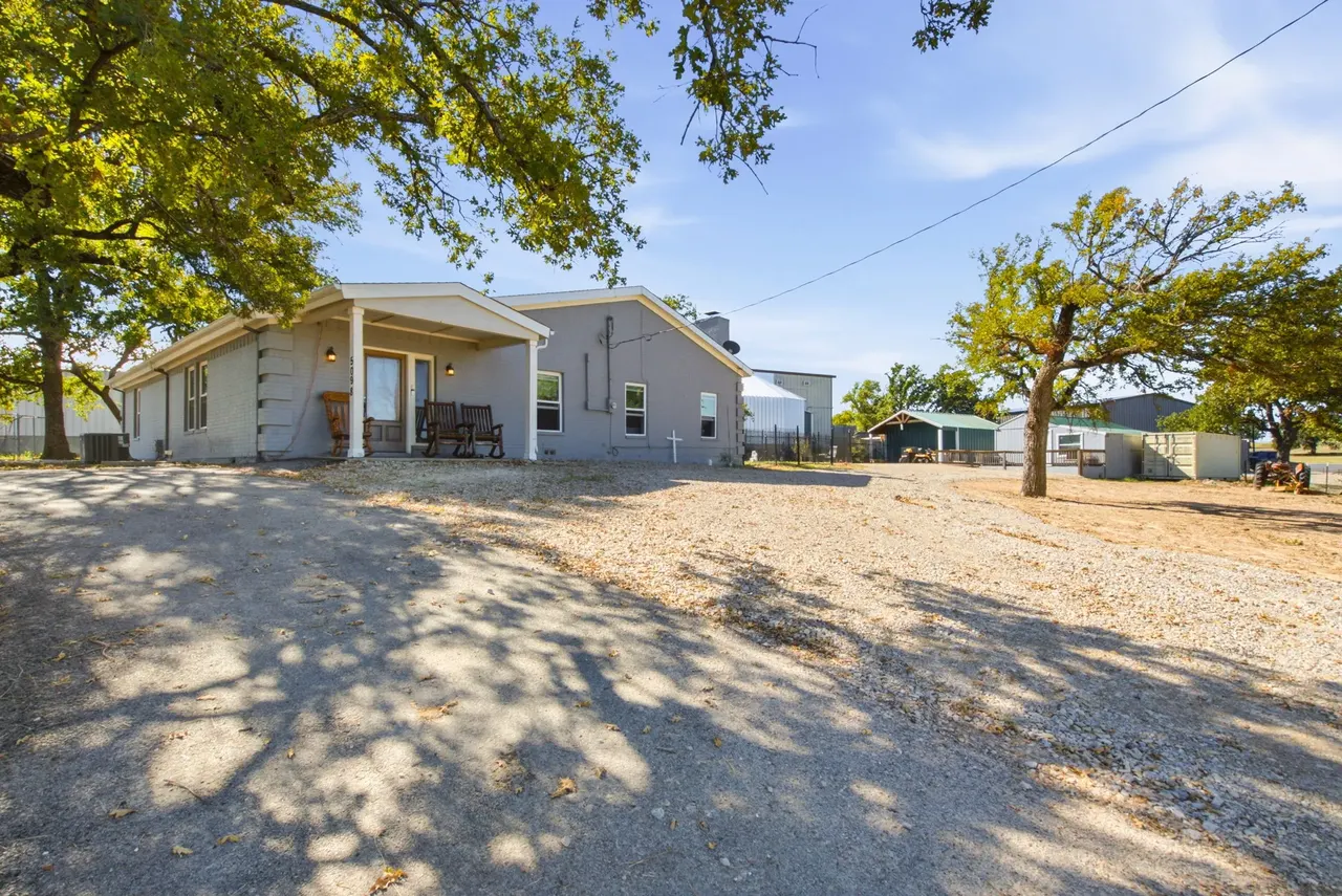 Gray single-story house with a covered front porch, behind a wide gravel drive under oak trees